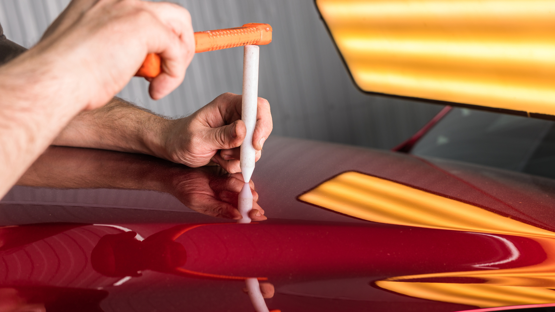 Hands using tool to remove dent from red car under a bright light.