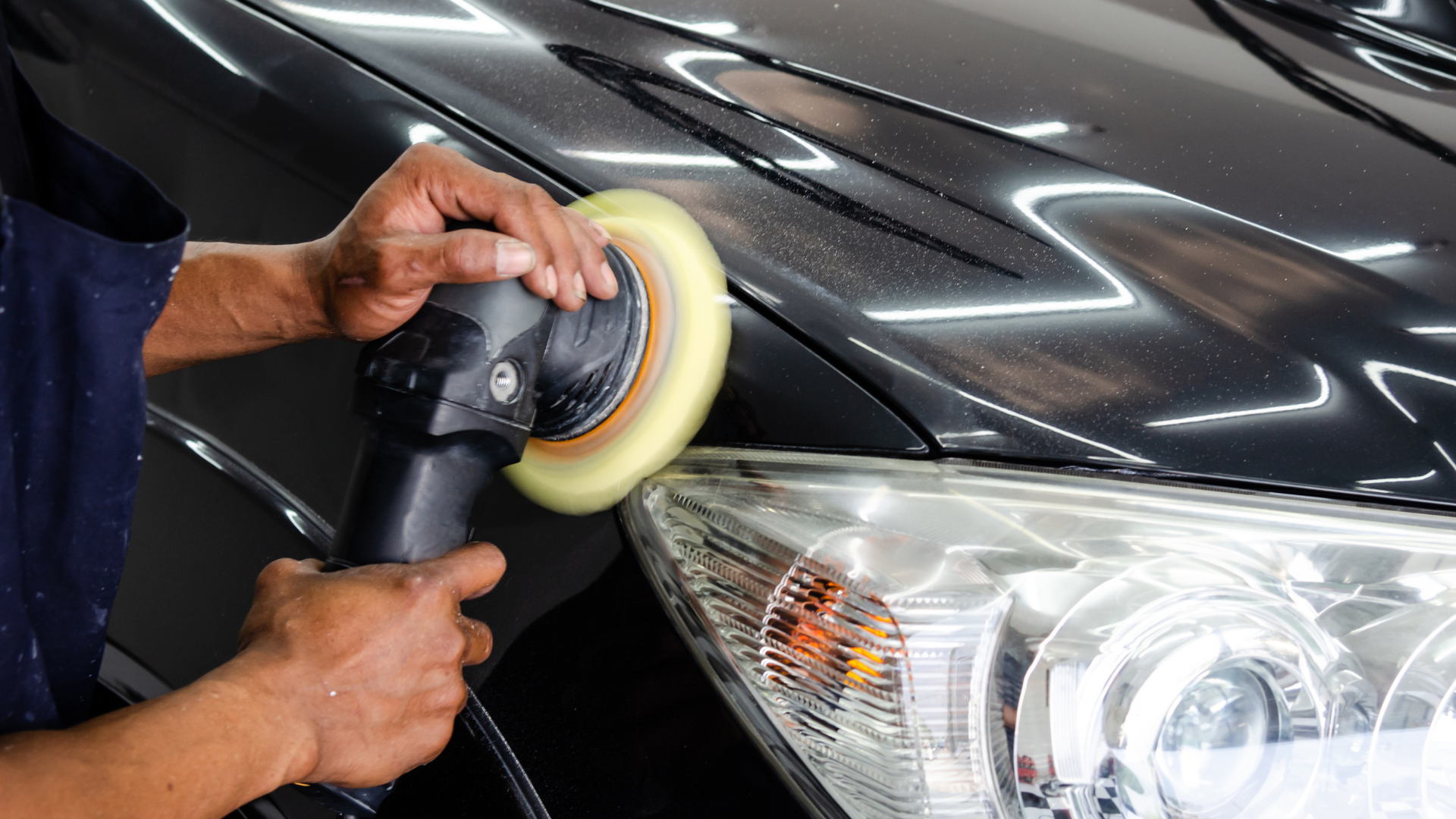 Person polishing a black car with a machine buffer; focus on the hood and headlight.