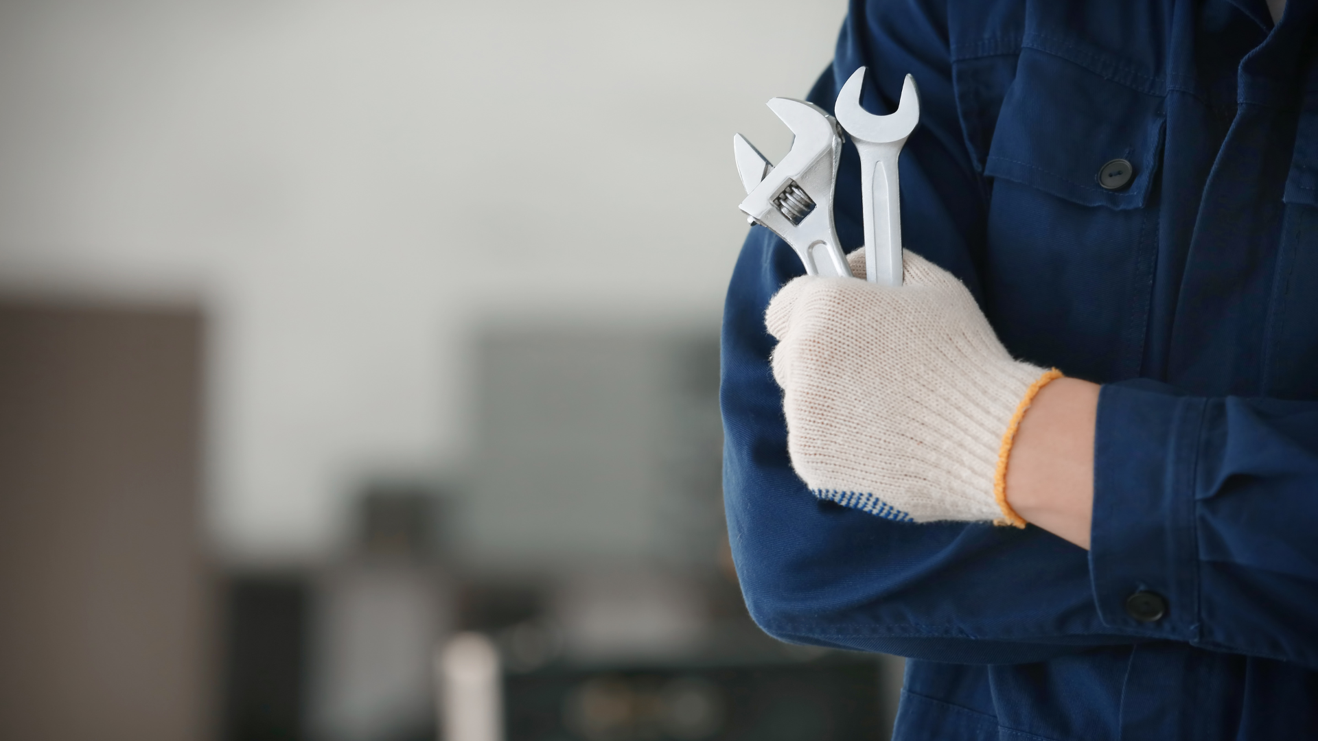 Mechanic holding wrenches, wearing gloves and a blue uniform; indoor setting.