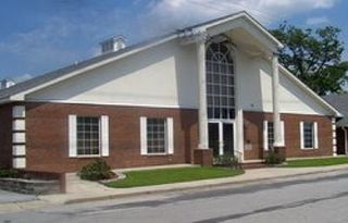 A one-story brick building with white trim, a large front arched window, and two white columns under a gabled roof.