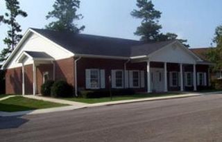 A brick office building with a dark shingled roof, white trim, and a front porch, surrounded by trees on a sunny day.