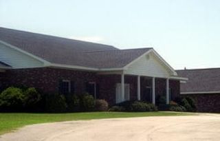 A low-slung brick building with a gabled porch entrance, white trim, and a shingled roof, surrounded by lawn and a dirt path.