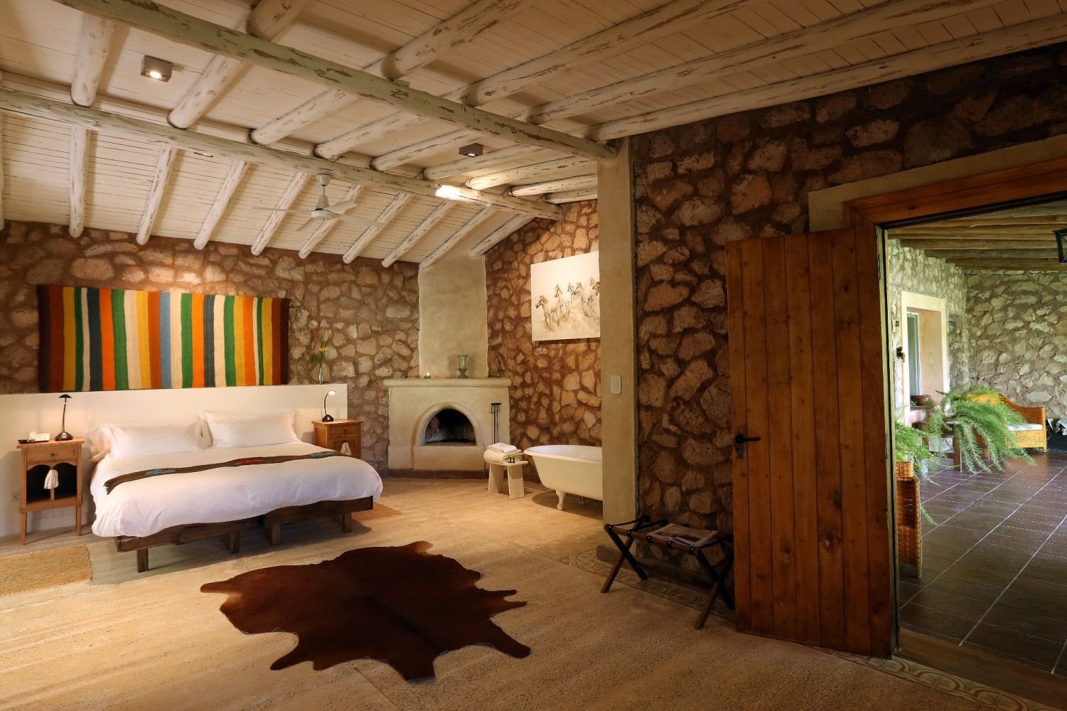 Bedroom with stone walls, white ceiling, bed with colorful textile, fireplace, and a door to an outdoor area.