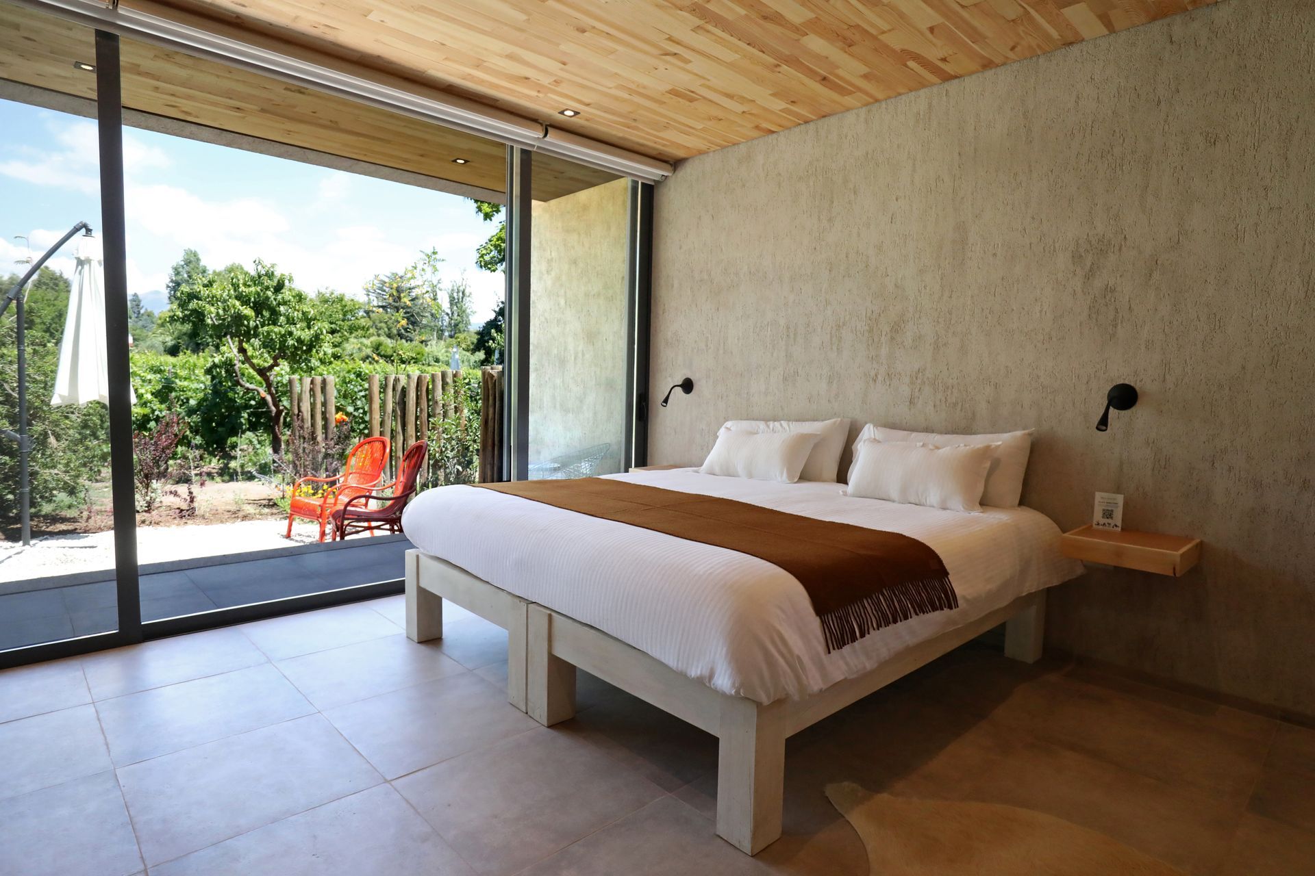 A modern bedroom with large windows overlooking greenery. A bed, rug, and wooden door are visible.