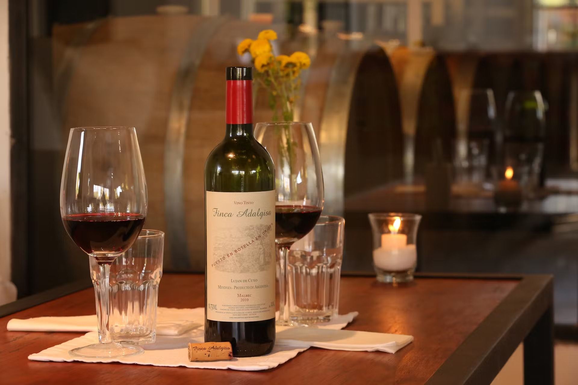 A wine bottle, glasses with red wine, and a candle on a table in front of barrels.