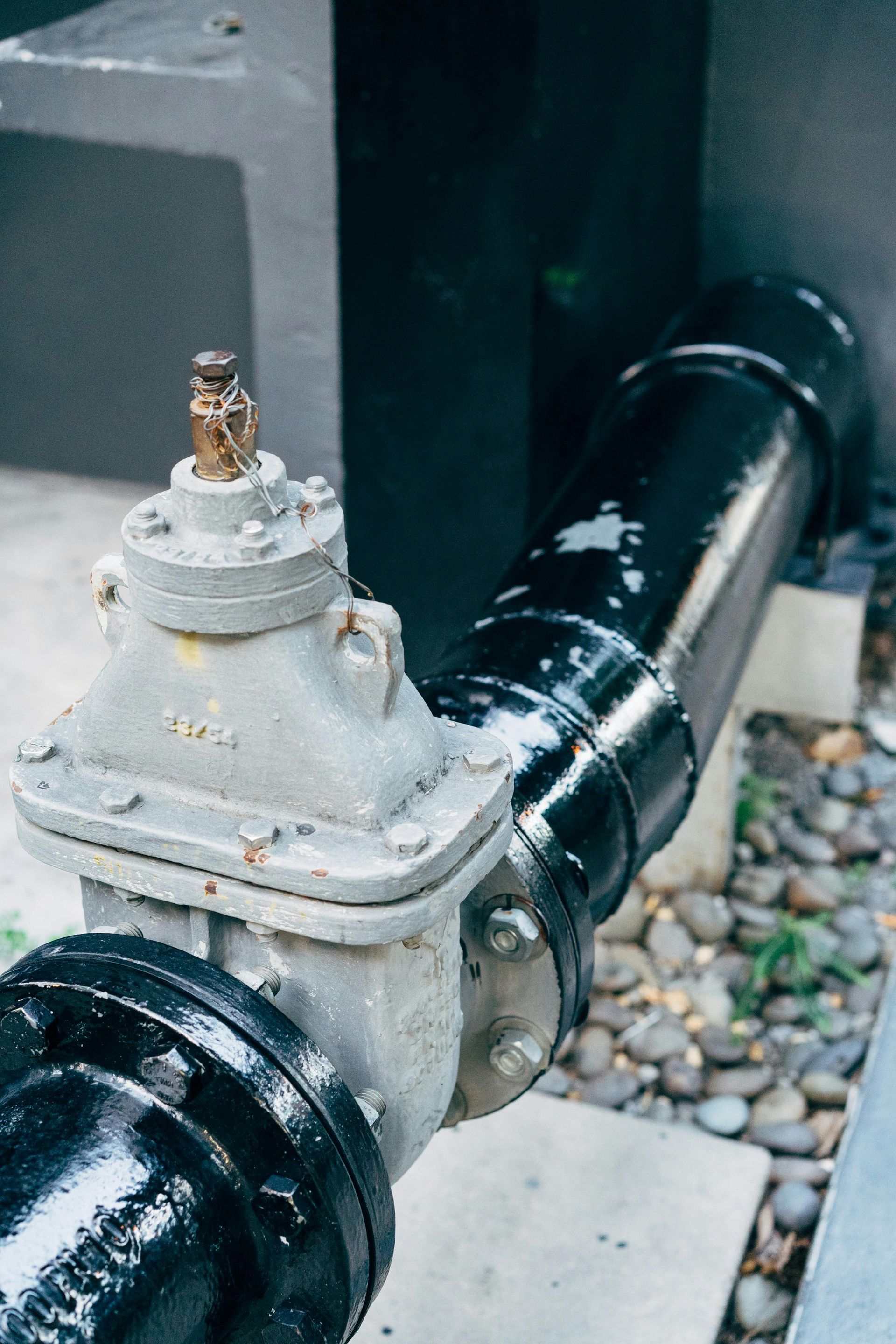 Black and silver industrial pipes and valve with a black wall in the background. Gravel and plants are at the bottom.