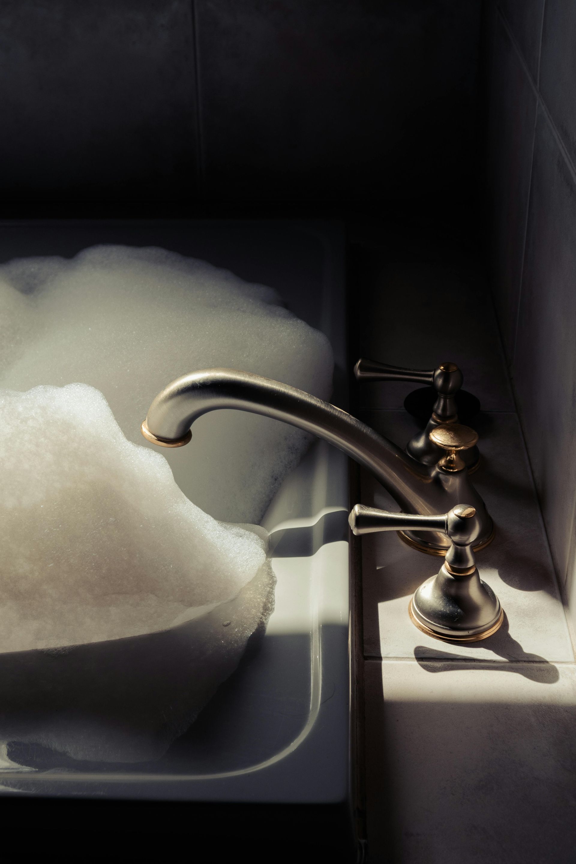 Bathtub filled with white suds, antique silver faucet with light and shadows.