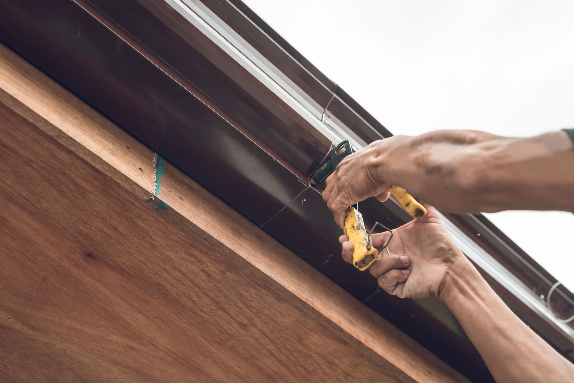 Person installing brown gutter with a yellow tool on a wooden building exterior.