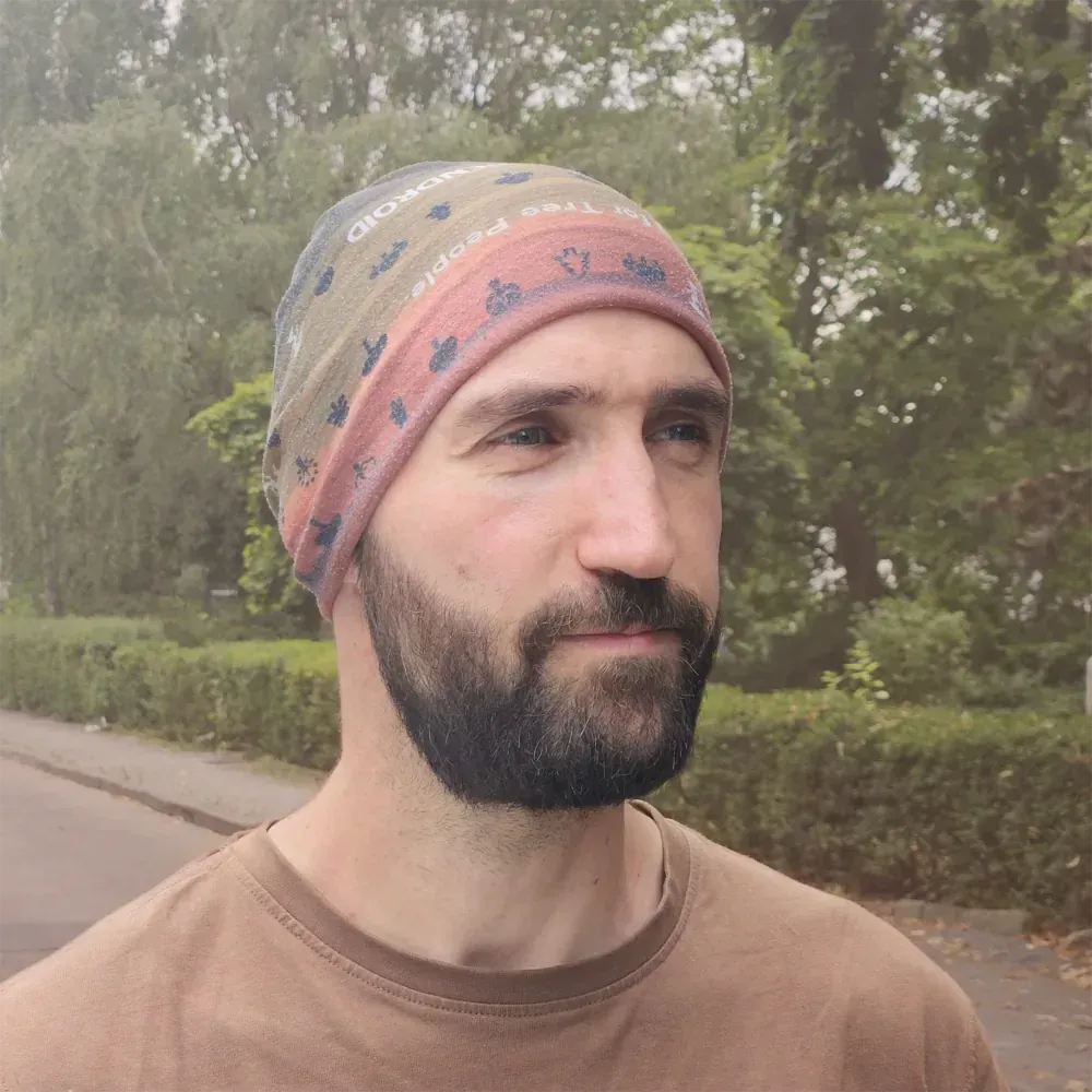 Mann mit Bart und gemusterter Mütze vor einem unscharfen Hintergrund mit Bäumen im Freien.