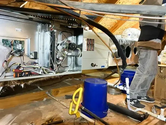 Person working on HVAC system in an attic. Blue bucket, electrical wires, insulation.