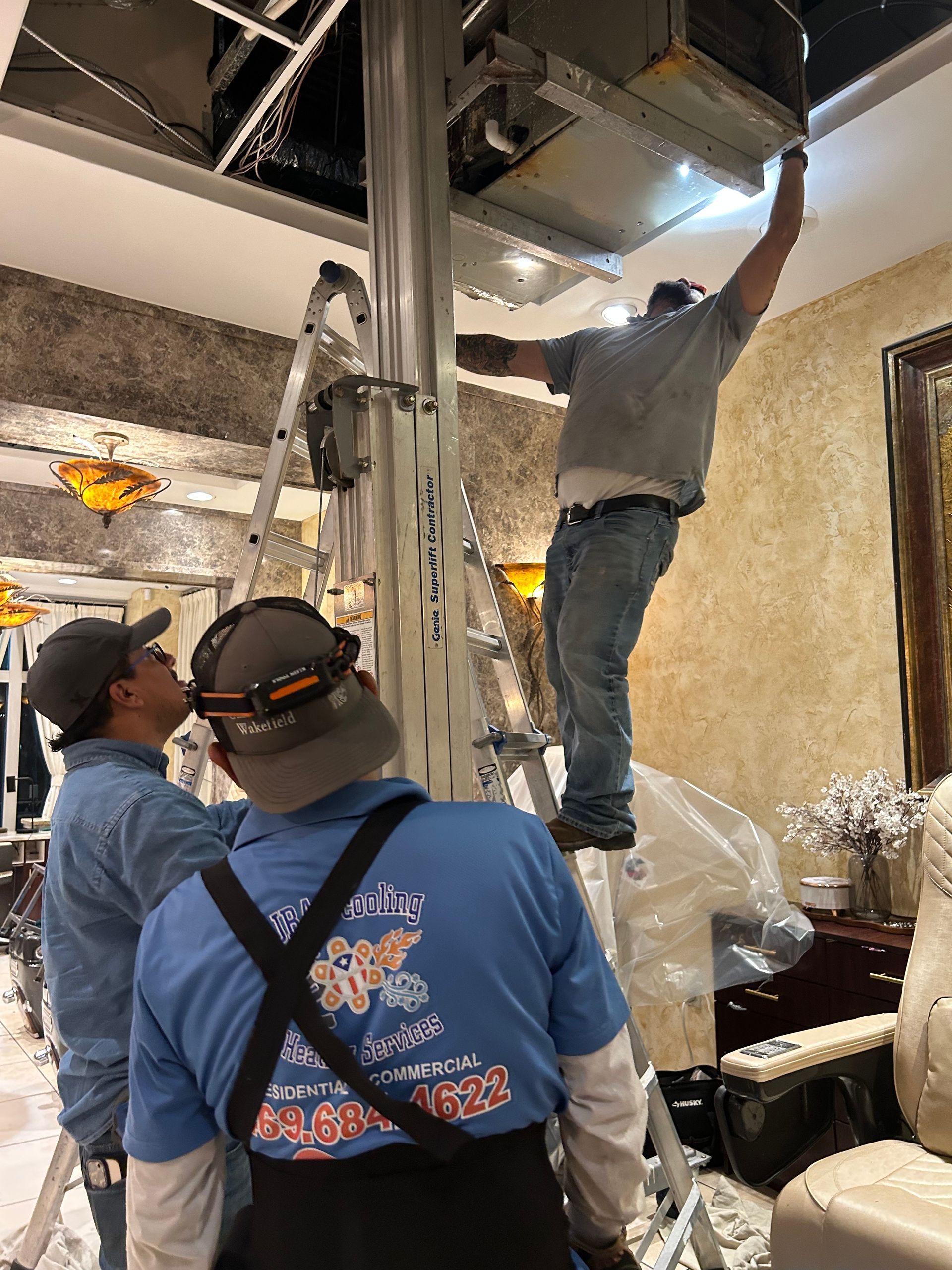 Three workers installing an air conditioning unit in a room with a damaged ceiling. One is on a lift, reaching up.