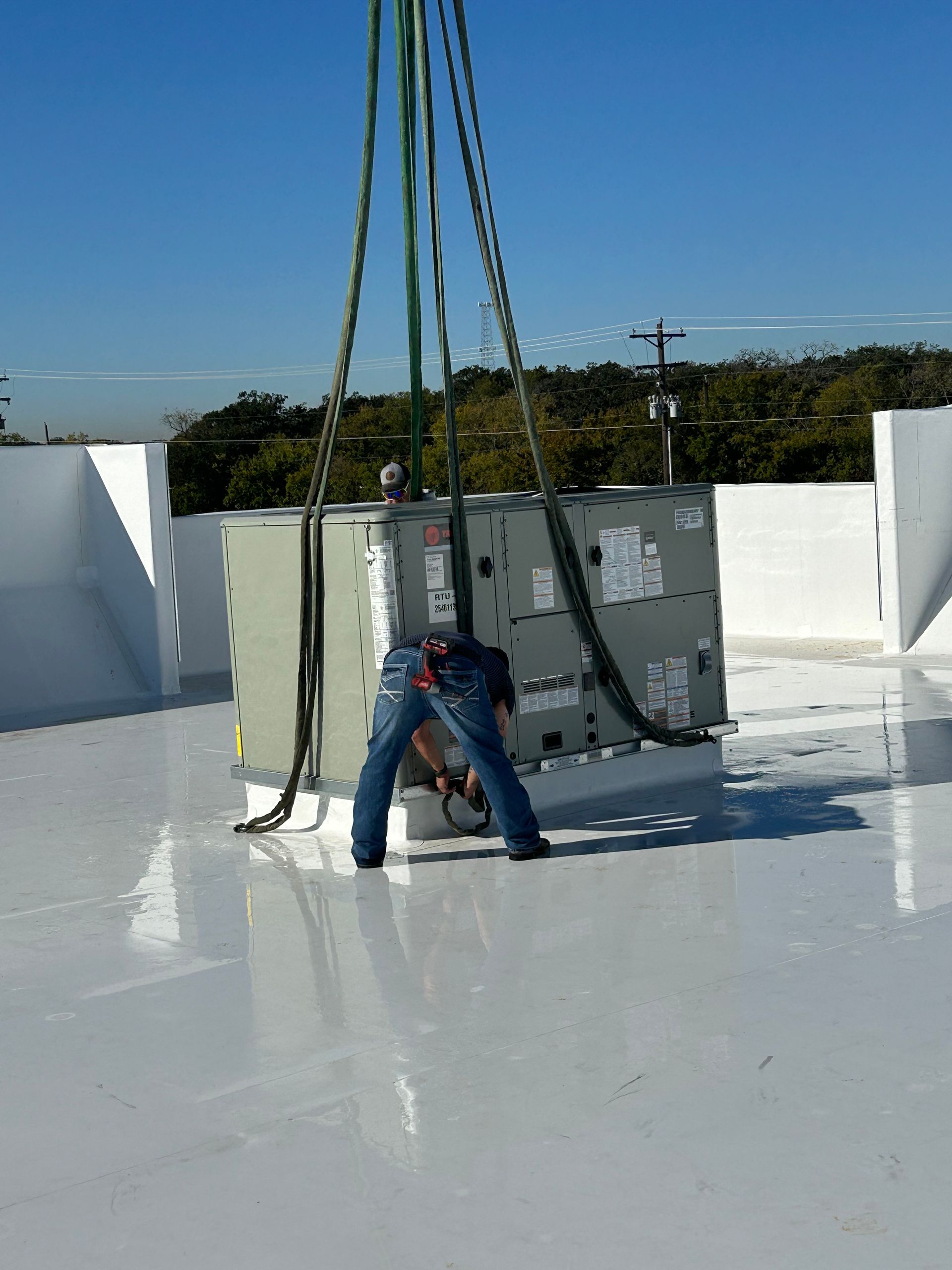 Person working on HVAC unit being lowered by a crane on a white rooftop.