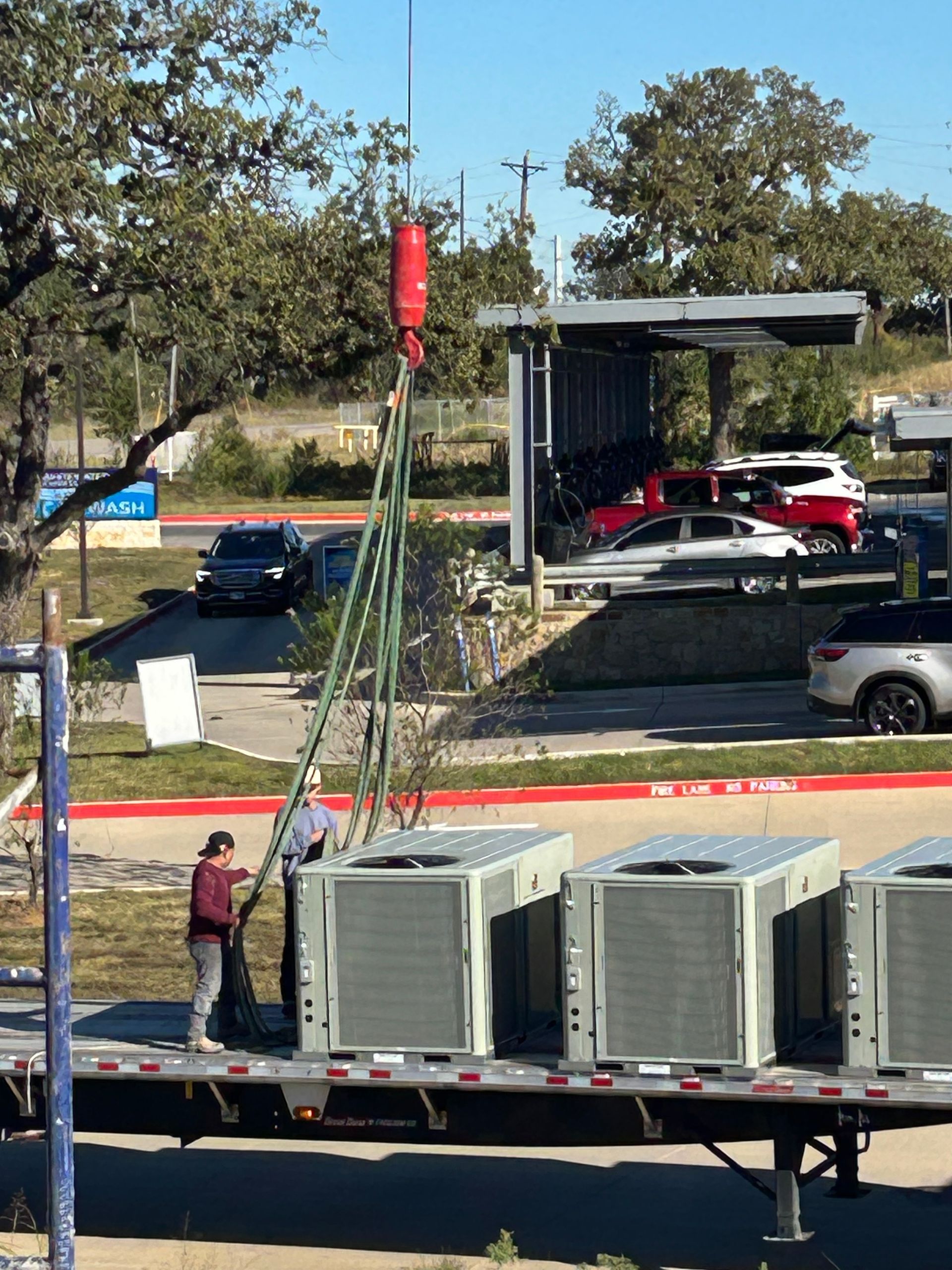 A crane lifting air conditioning units from a truck. Workers secure the units on a trailer, outside, on a sunny day.
