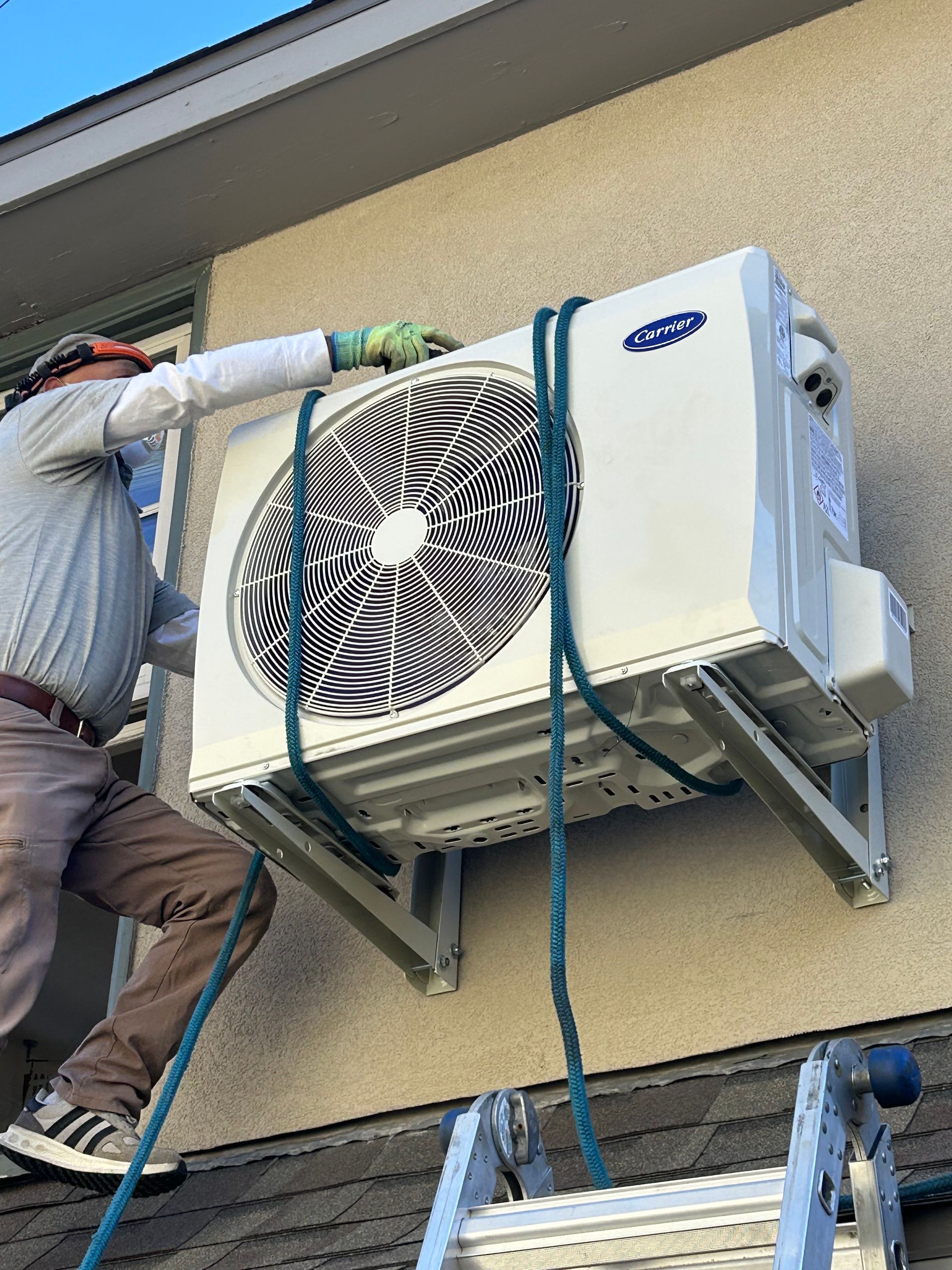 Man installing an air conditioner unit on a building exterior, secured with ropes.