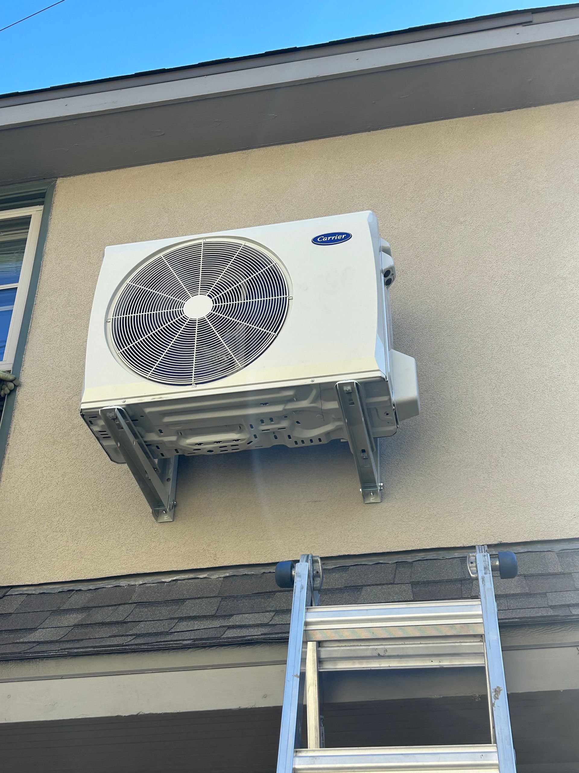 White Carrier AC unit mounted on a stucco wall above a ladder, blue sky in background.