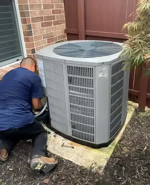 Man working on an outdoor air conditioning unit. Unit is gray, on a concrete pad, near a brick wall and a fence.