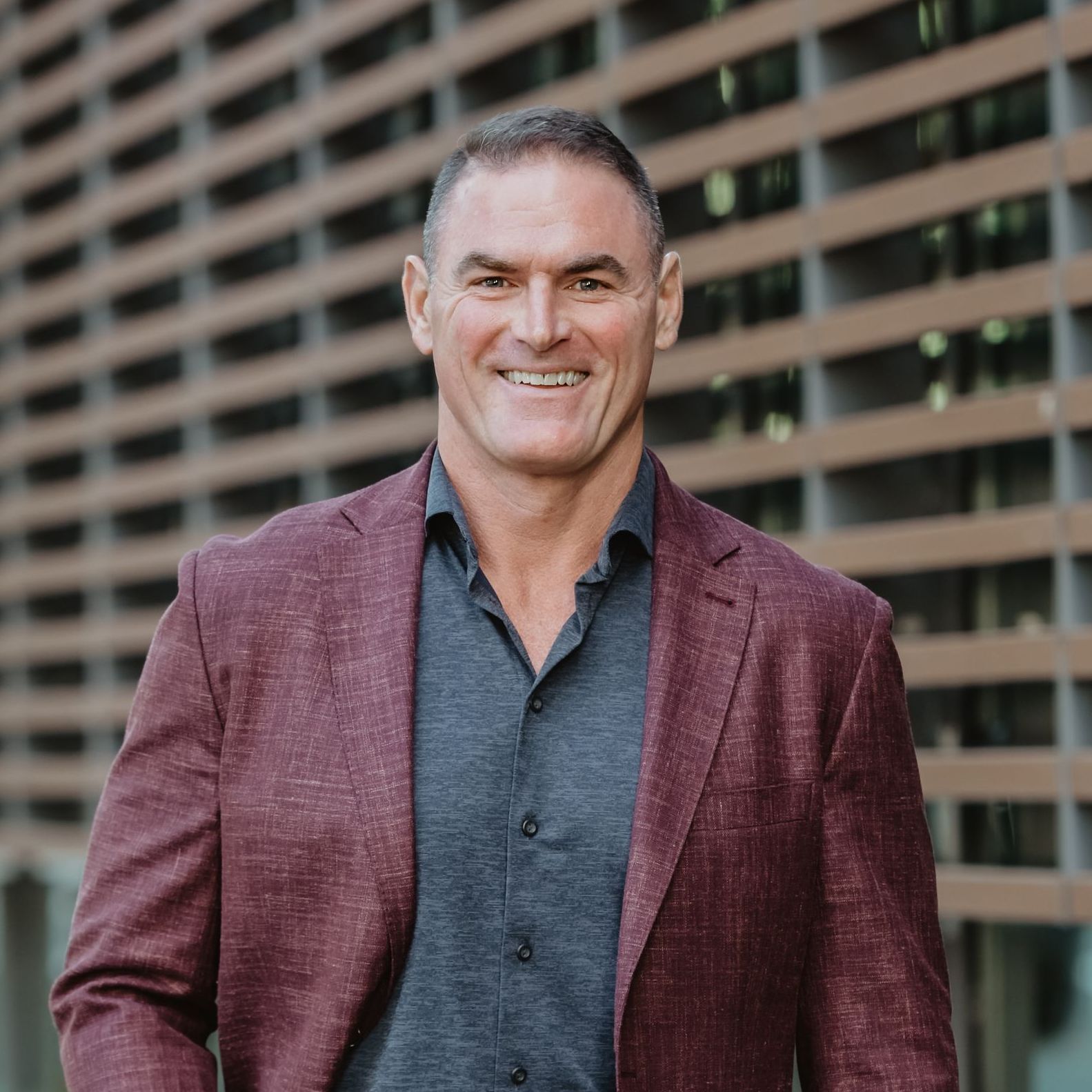 A man in a maroon jacket and gray shirt is standing in front of a building.