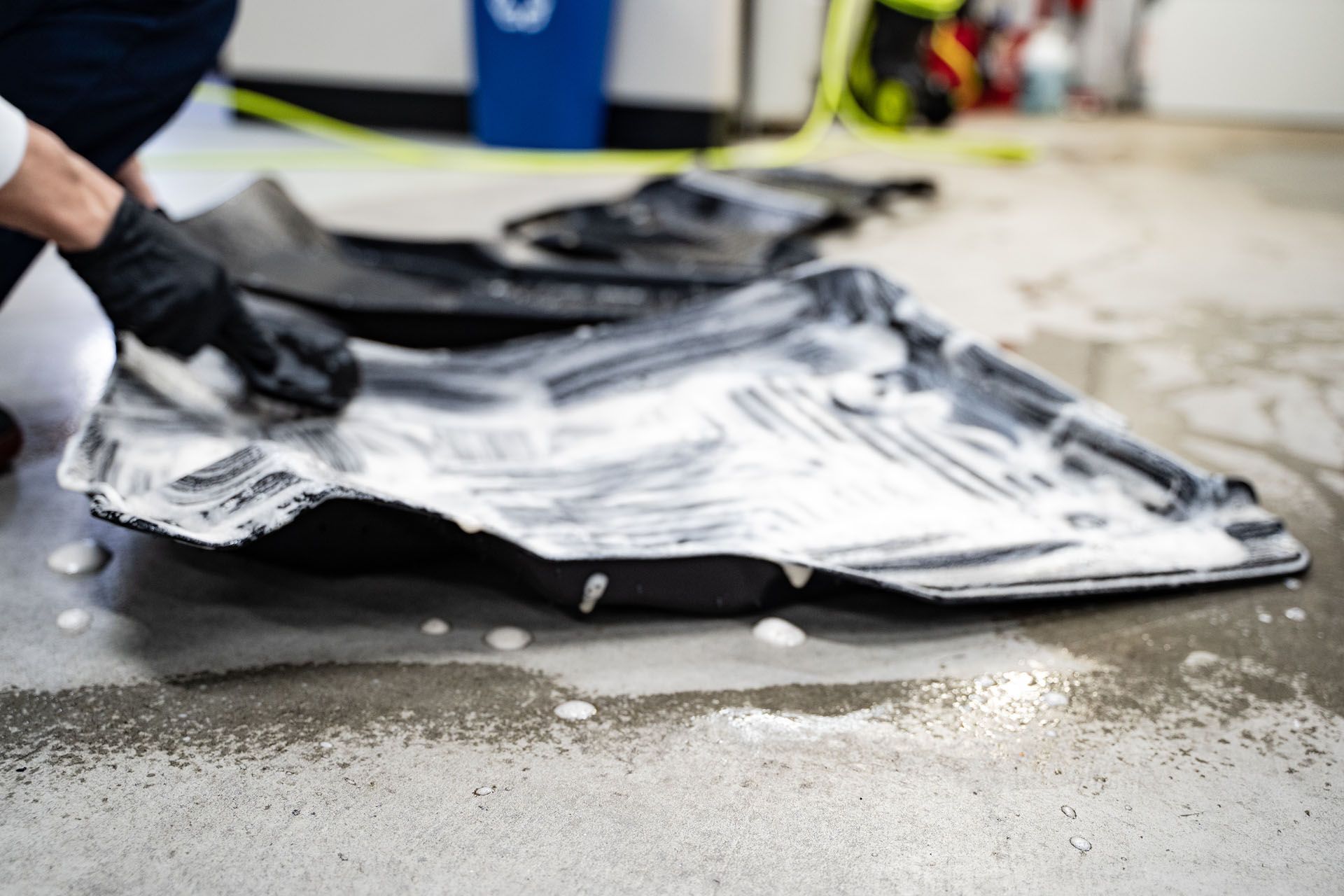 Person in black gloves scrubbing a car floor mat with soapy water on a concrete floor.