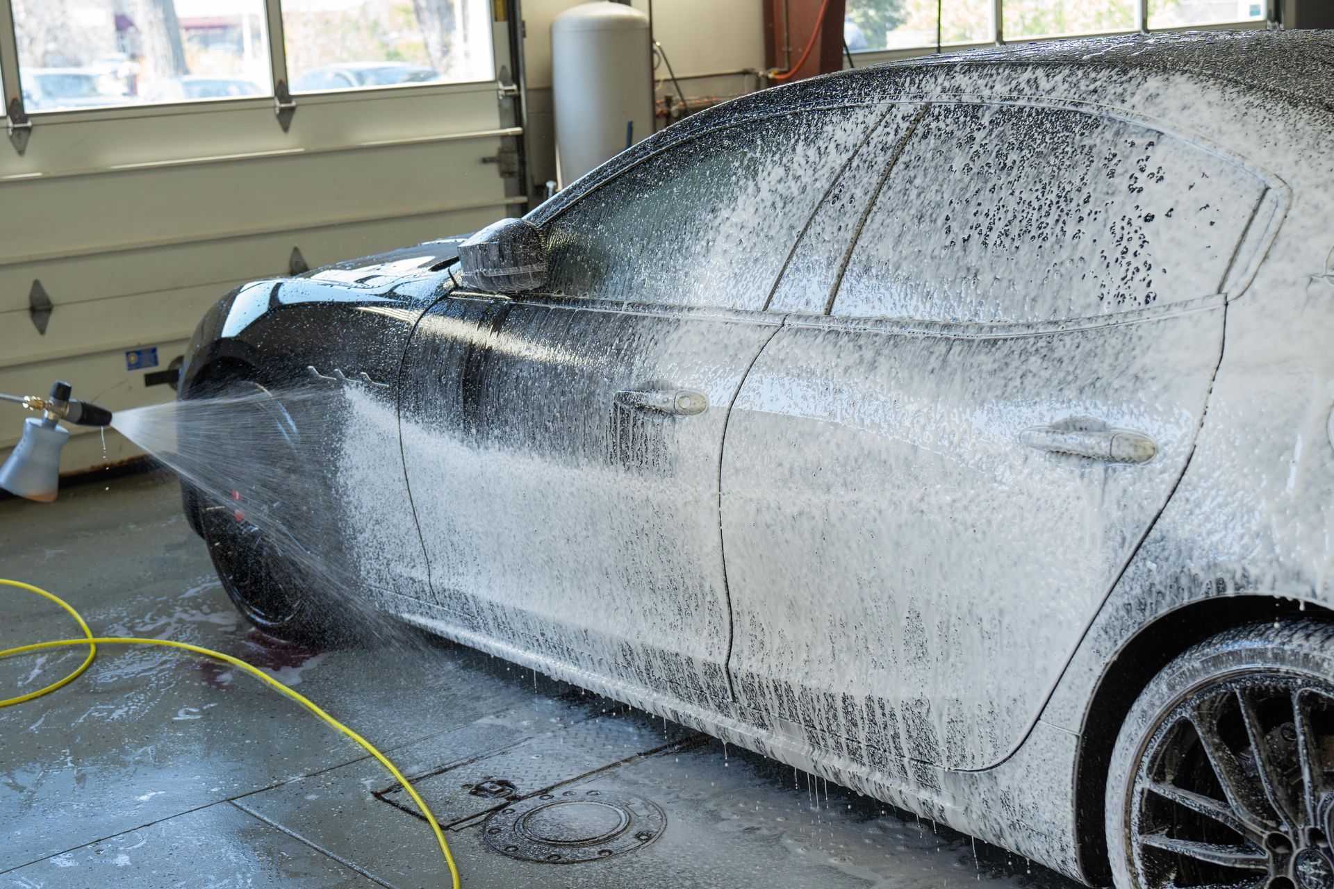 Black car covered in white foam, being washed in a garage setting. A yellow hose sprays the side of the car.