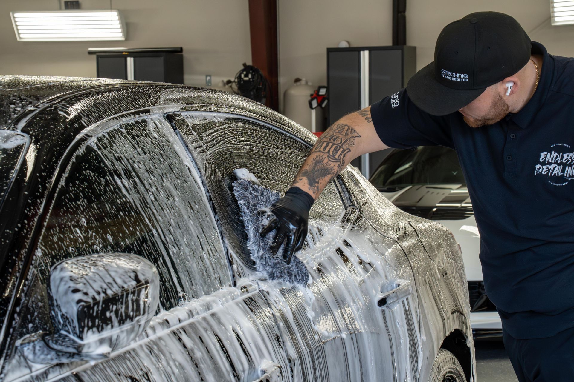 Man in a cap and gloves washing a black car with soapy water in a garage.