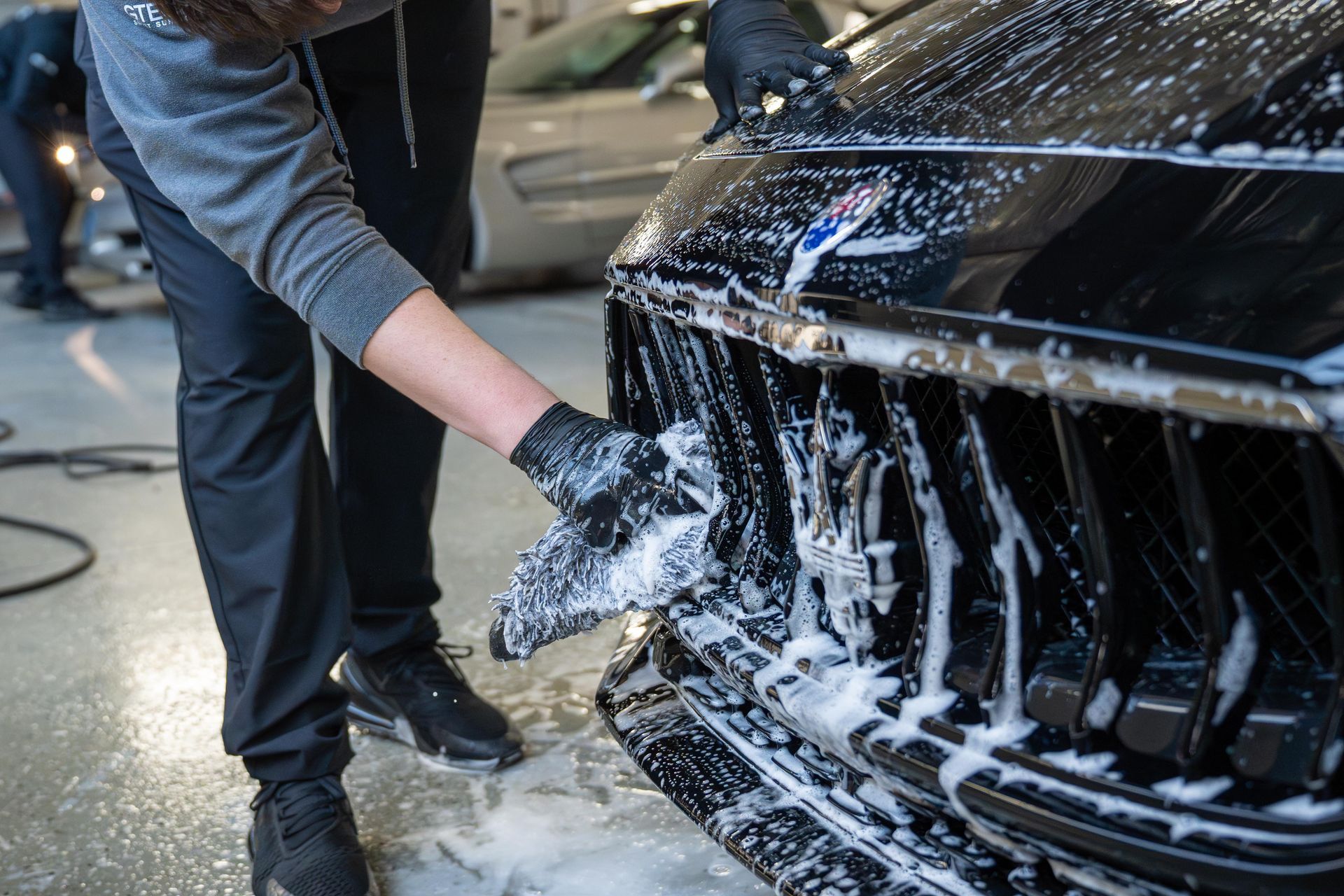 Person in black gloves washing a black car's grill with soapy foam in a garage.