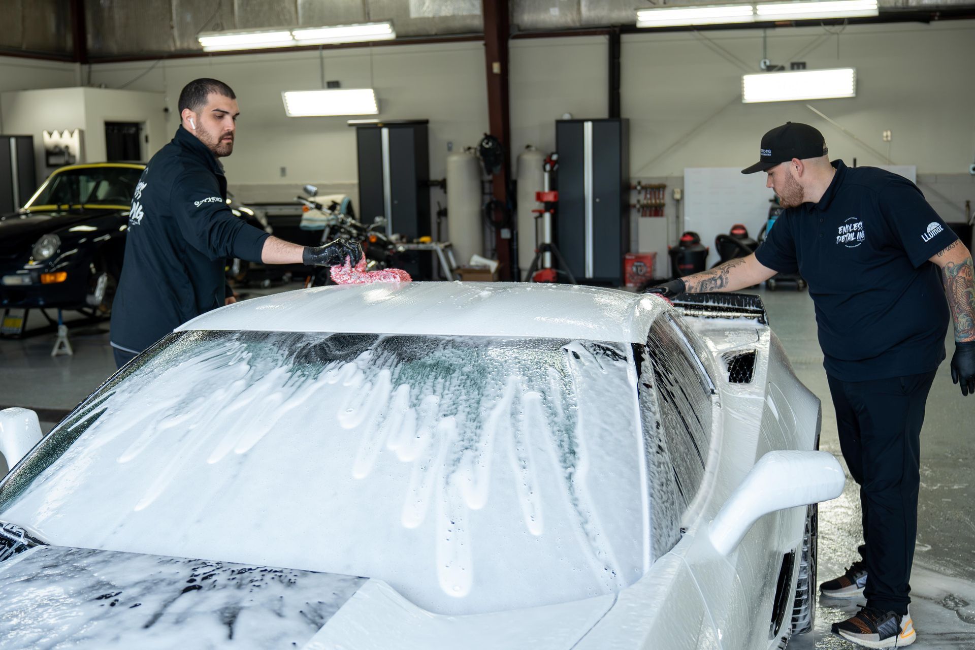 Two men washing a white sports car in a well-lit garage. They are applying soapy foam to the car's surface.