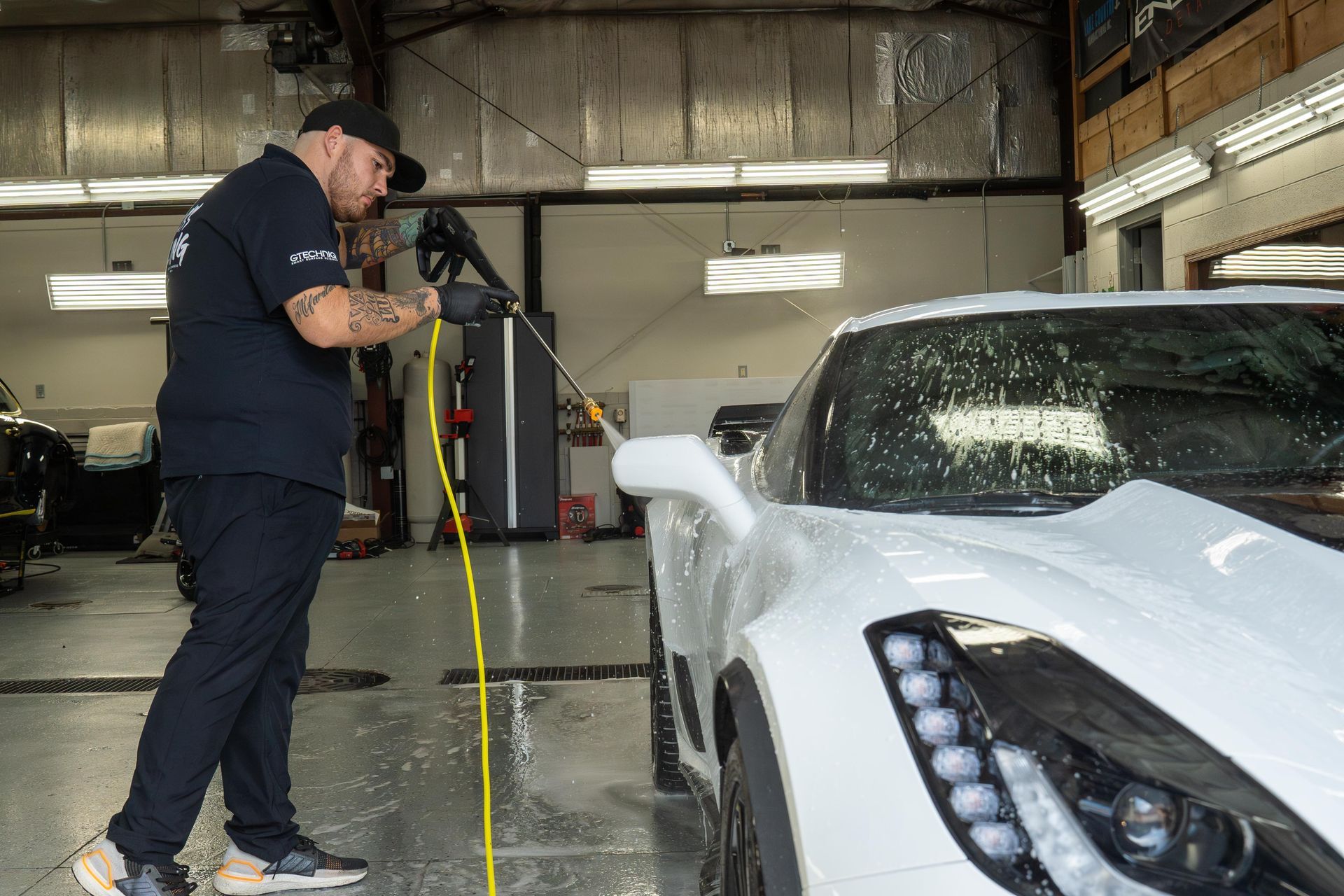 A person wearing black gloves and a hat washes a white sports car with a pressure washer inside a garage.