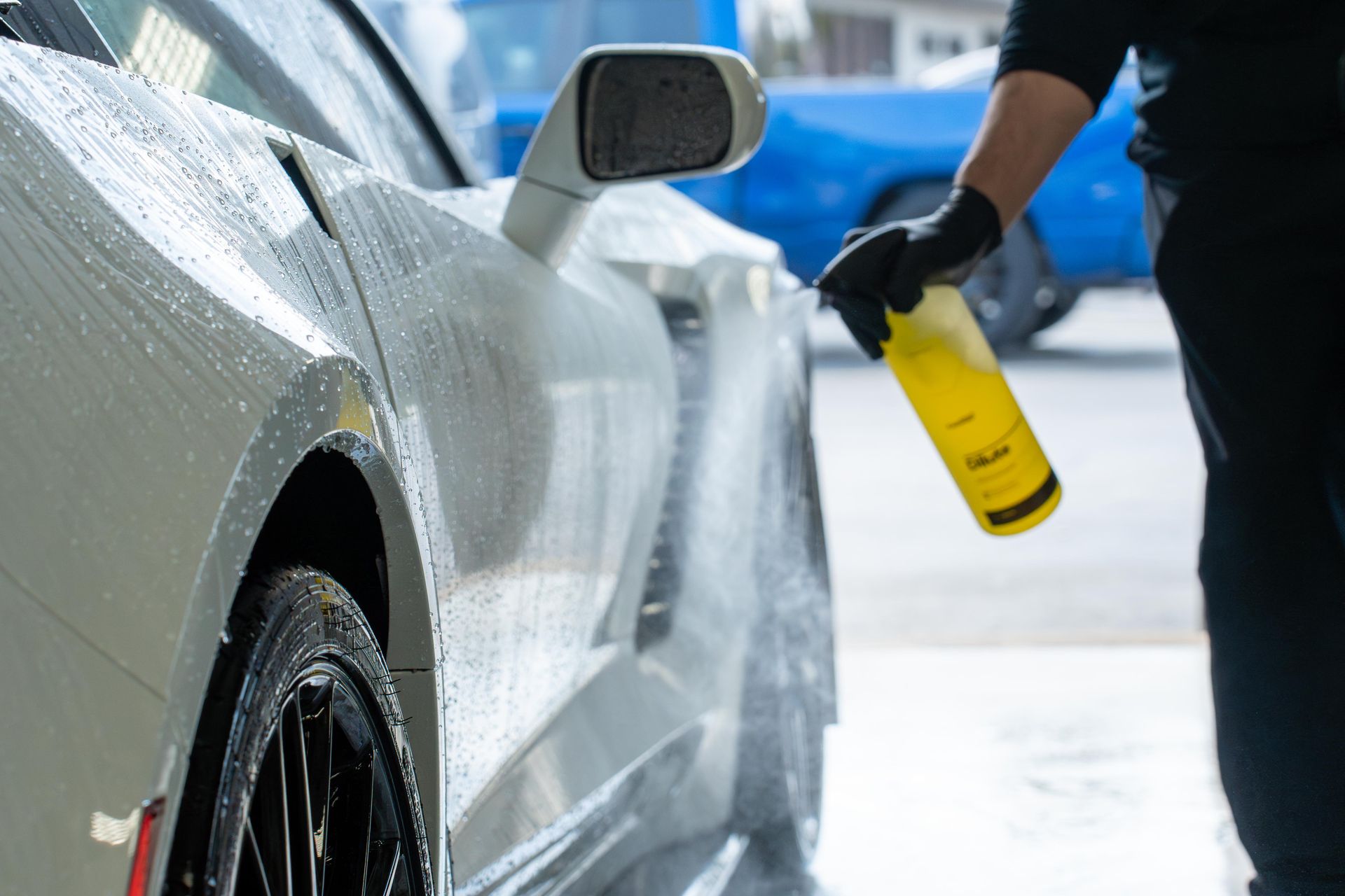 Person washing a white sports car with a yellow spray bottle, outdoors.