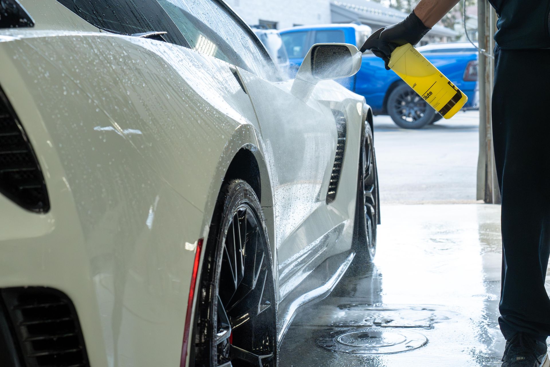 A person in black gloves sprays a white sports car with a yellow bottle at a car wash.