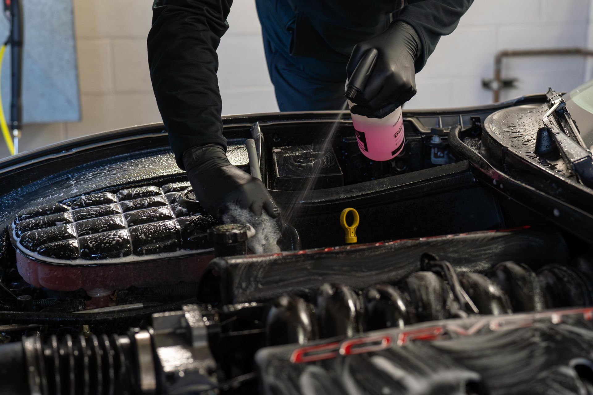 Person wearing black gloves spraying and scrubbing a car engine with soapy water.