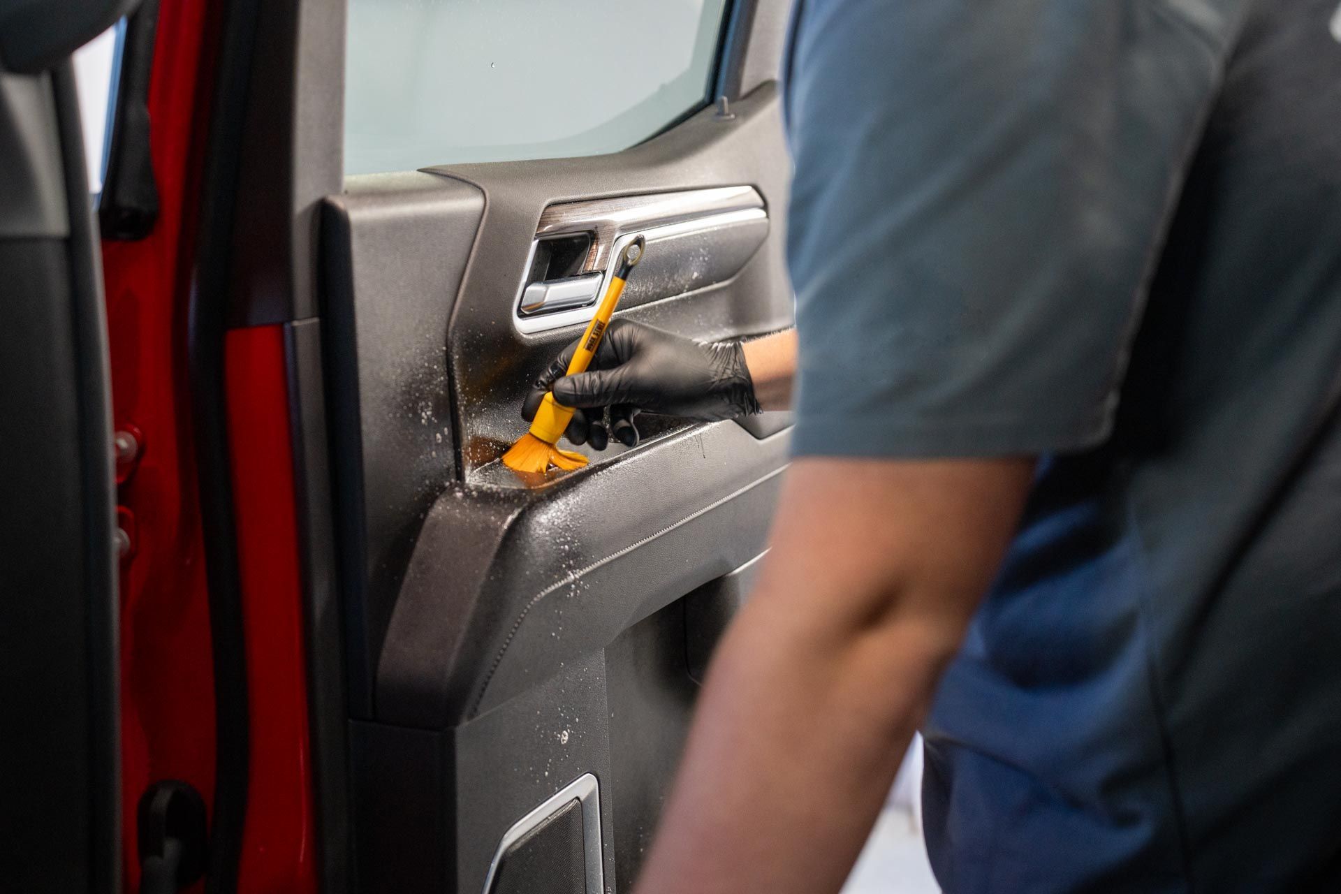 Person cleaning the interior door panel of a red car with a brush. They wear a black glove, and the panel is wet with cleaning solution.