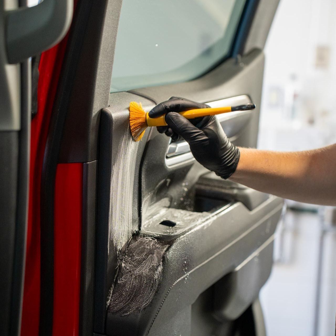Person wearing a black glove scrubs a car door panel with a brush. The door is red, the setting is indoors.
