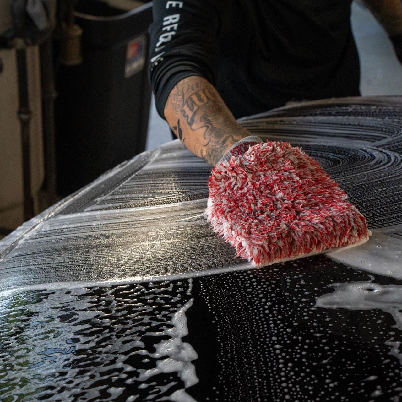 A person with tattoos washes a car with a red wash mitt covered in soapy foam.