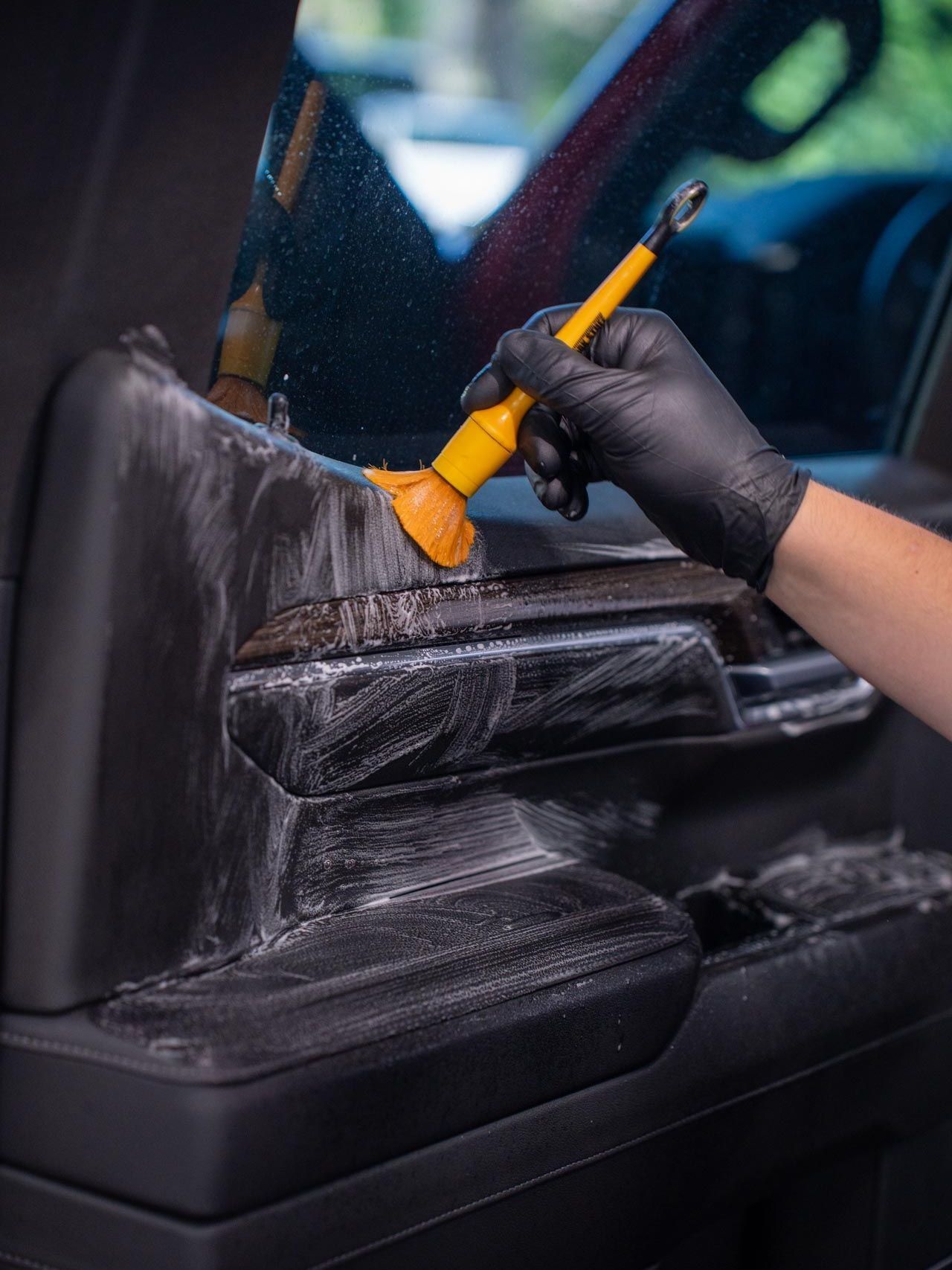Person wearing black gloves cleaning a car's interior with a brush. The car's door panel is black with cleaning solution applied.