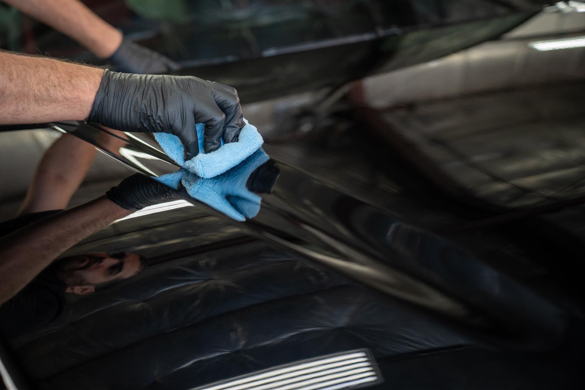 Hands in black gloves wiping a shiny black car surface with a blue microfiber cloth.