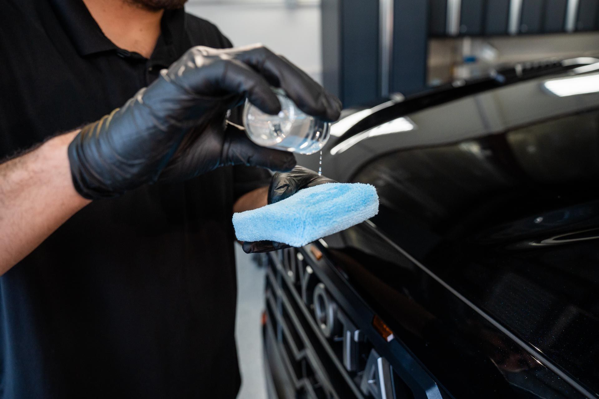 Person wearing black gloves applying liquid to a car's black surface with a blue applicator in a garage setting.