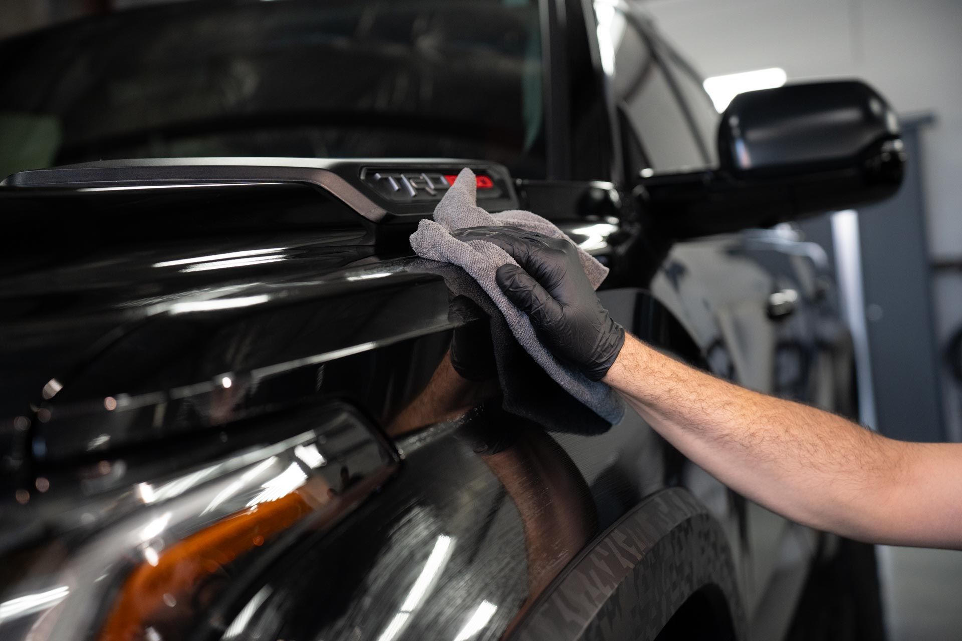 A person wearing a black glove wipes a black car's hood with a gray cloth.