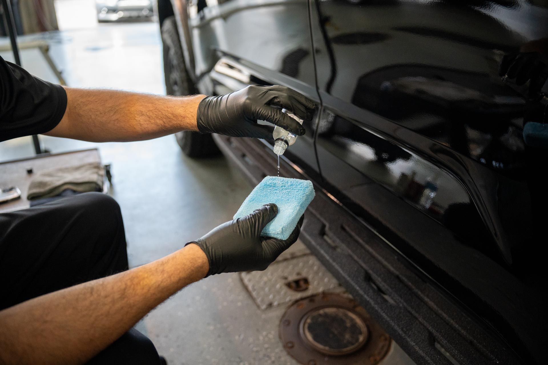 Person wearing black gloves applying a product to a black vehicle's side panel with a sponge.