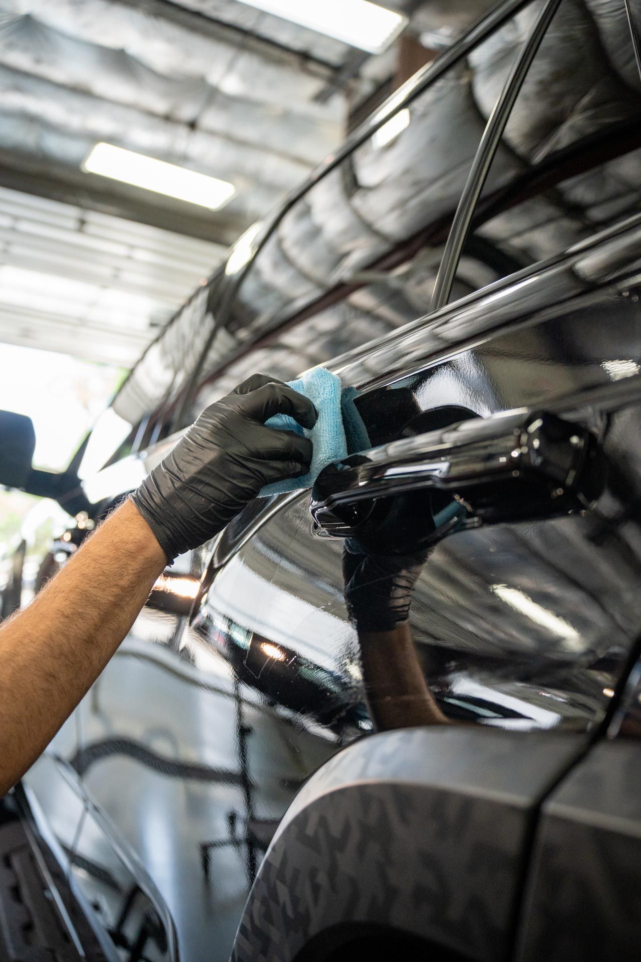 A person wearing black gloves is polishing a black car in a well-lit garage, using a blue microfiber cloth.