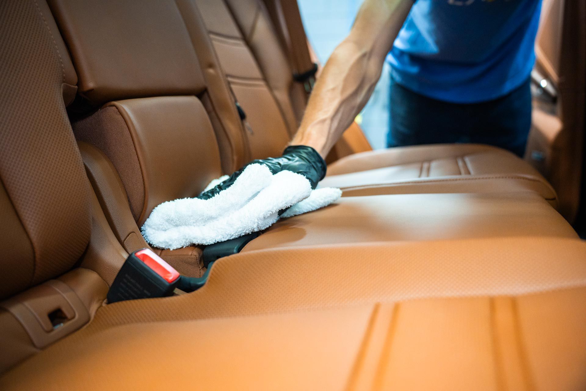 Person cleaning a tan leather car seat with a white microfiber cloth.