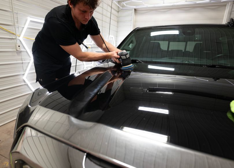 A man polishing a black car hood indoors. He is using a machine buffer, focusing on the glossy surface.