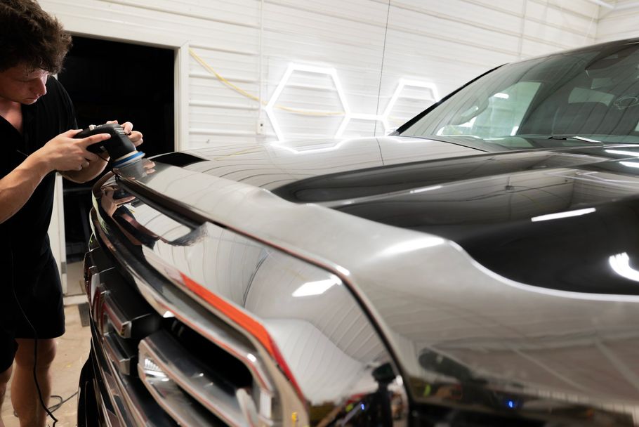 A person polishing a black pickup truck's hood in a bright garage. Chrome bumper gleams.