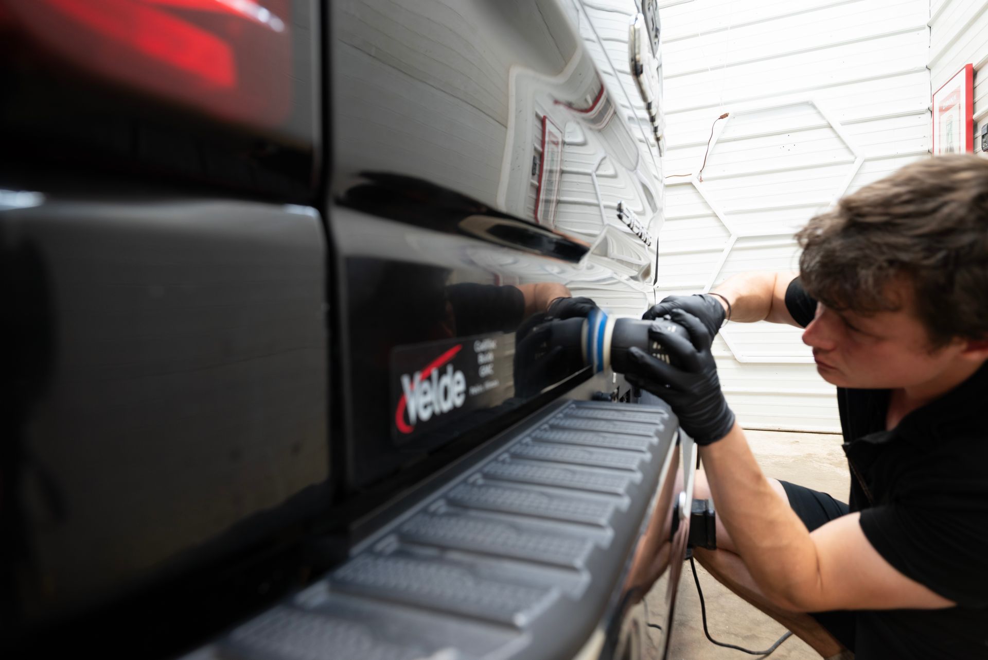 Man polishes a black car bumper with a power polisher. He wears black gloves and a black shirt. The setting appears to be a garage.