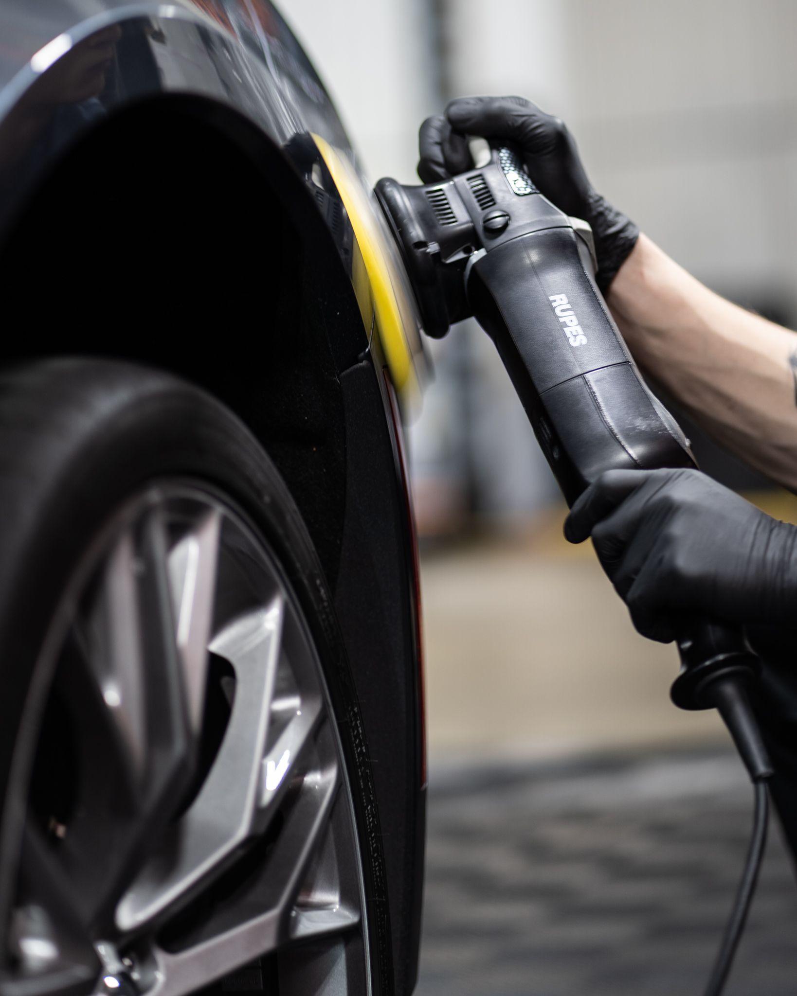 A person polishing a black truck in a well-lit garage, using an electric buffer.