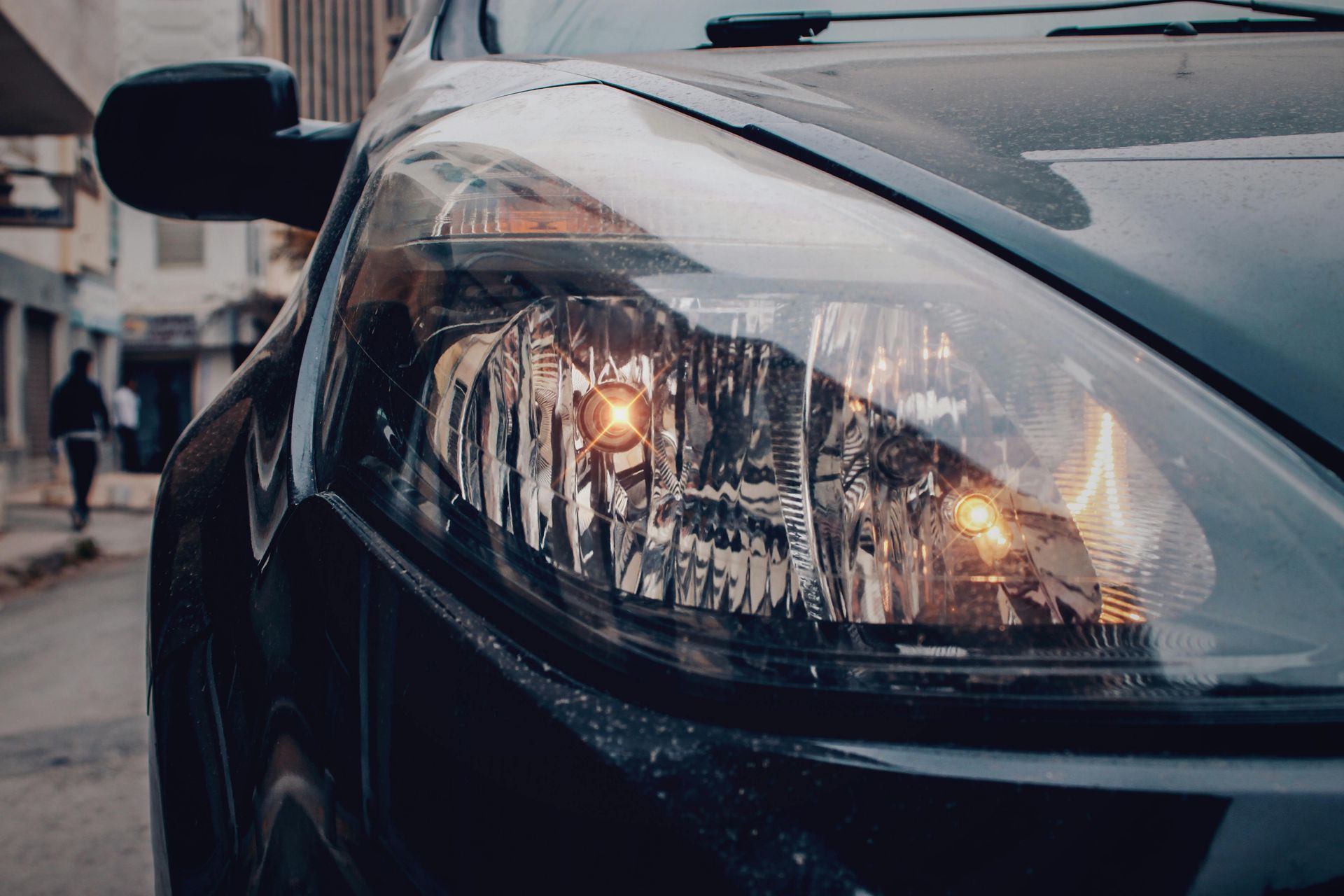 Close-up of a black car headlight, reflecting warm light. The car is parked on a narrow street, with a blurry pedestrian in the background.