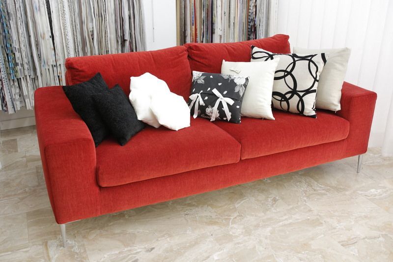 Red velvet sofa with multiple decorative pillows on a tiled floor. Fabric samples in background.