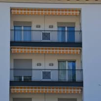 Three-story building with balconies, awnings, and glass railings. The facade is white.