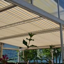 Beige slatted awning over an outdoor seating area. Plants in the foreground, trees and water visible in the background.