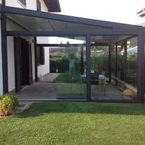 Glass-enclosed sunroom with a black frame and roof, next to a grassy lawn.
