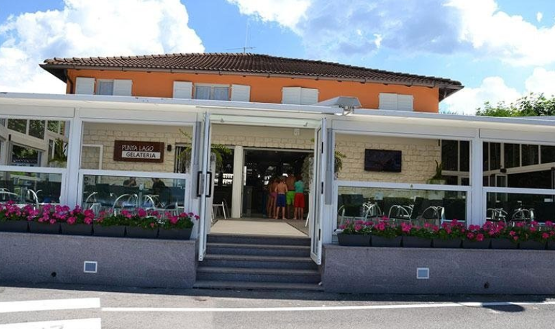 Restaurant with glass walls and a tiled roof, people inside, pink flowers in window boxes, sunny day.